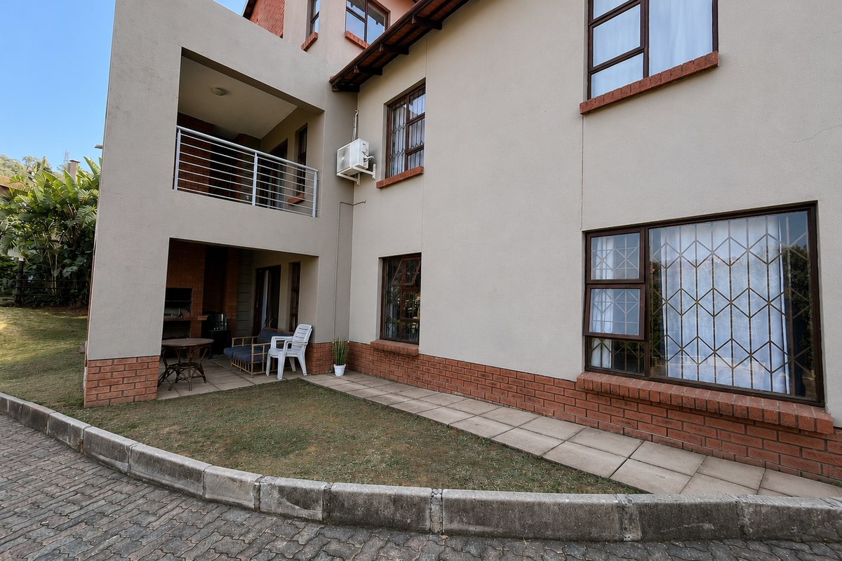 Rear exterior view of the apartment building featuring a covered patio with a built-in braai, balcony, and paved garden area.