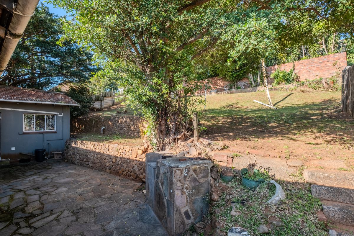 Rear garden area featuring a stone-paved patio, built-in stone braai, and tiered landscaping with brick boundary walls.