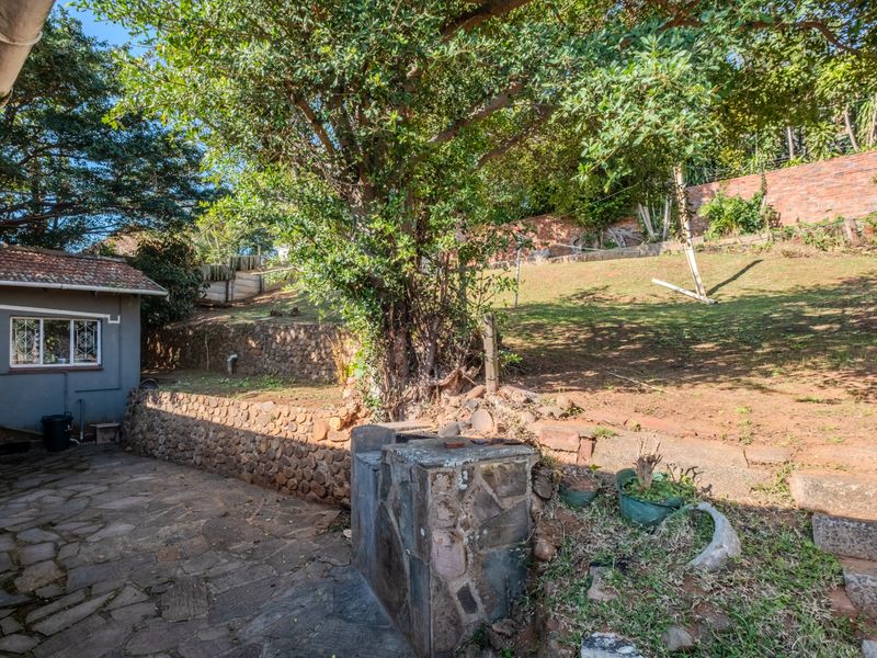 Rear garden area featuring a stone-paved patio, built-in stone braai, and tiered landscaping with brick boundary walls.