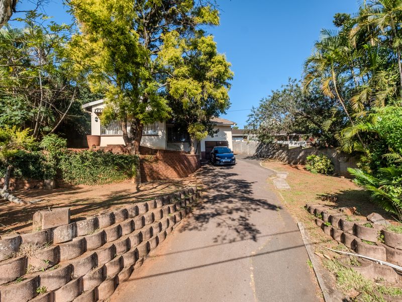 Front exterior view of the property featuring a long asphalt driveway, brick retaining walls, and mature landscaping leading to the main entrance and garage.
