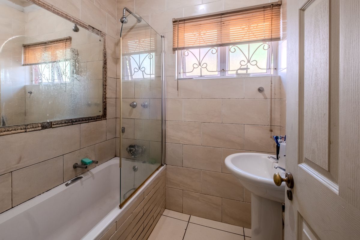 A dated bathroom featuring a pedestal basin, bathtub with a glass shower screen, and neutral wall tiling. Requires cosmetic updates to modernize the space.