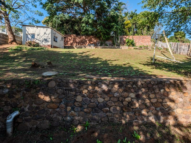 A sloped backyard garden featuring a stone retaining wall, mature trees, and brick boundary walls, offering potential for landscaping and outdoor development.
