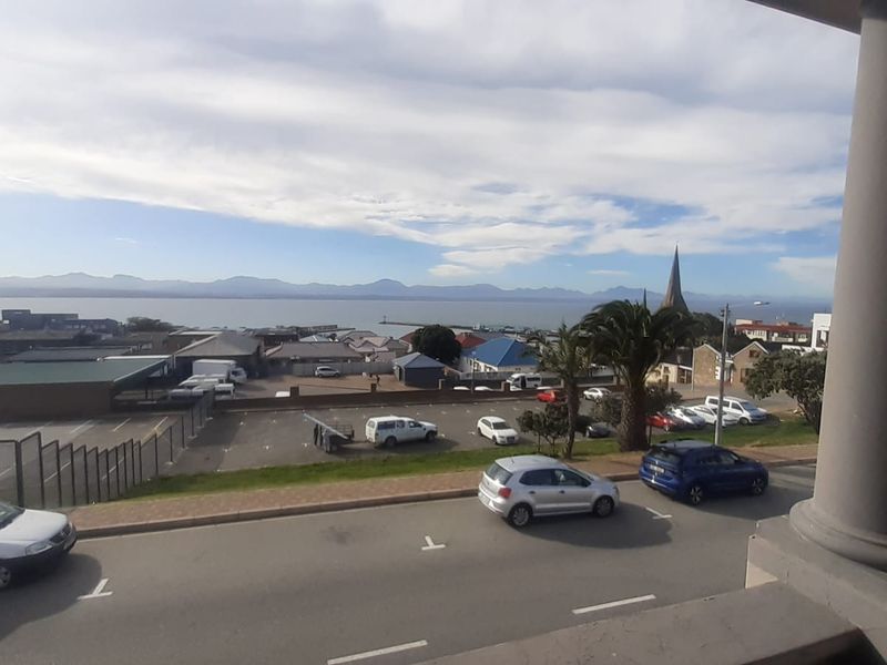 Elevated coastal view overlooking the street, parking area, and distant mountains from a balcony.