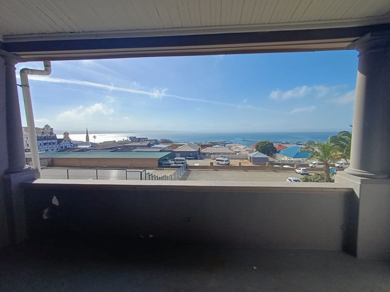 A covered balcony featuring a concrete balustrade and columns, offering an expansive elevated view of the surrounding town and coastline.