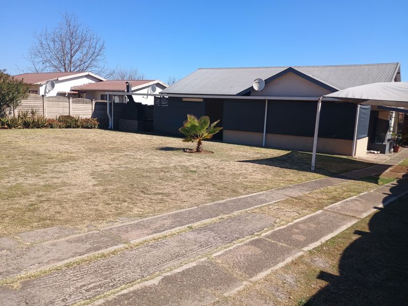 Rear view of the property featuring a spacious lawn, concrete driveway, and a covered patio area with a corrugated metal roof.