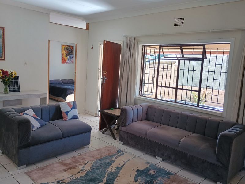 A living room featuring tiled flooring, large windows with security bars, and direct access to an adjacent bedroom.