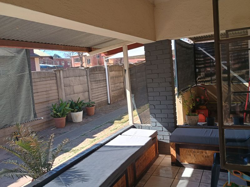 A covered outdoor patio area featuring tiled flooring, a painted brick support pillar, and a corrugated metal roof, overlooking a private garden space.