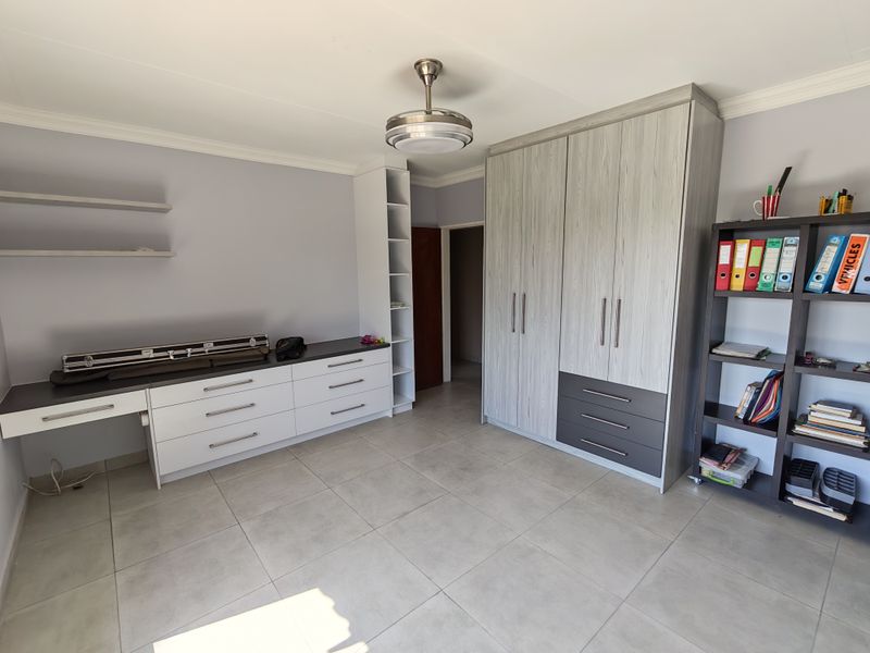A well-maintained bedroom featuring modern built-in cupboards, a functional built-in desk, floating shelves, and neutral tiled flooring.