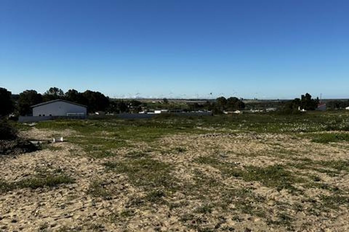A view of a vacant residential plot of land with sandy terrain and a boundary wall in the distance.