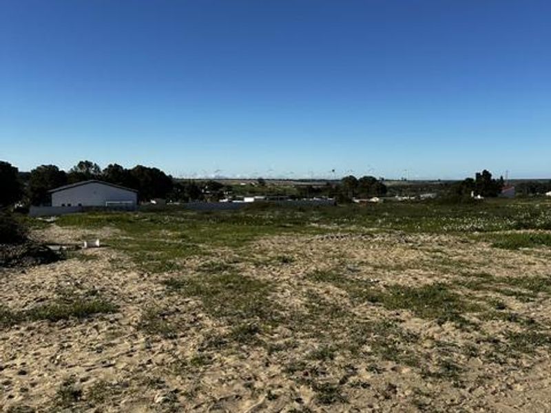 A view of a vacant residential plot of land with sandy terrain and a boundary wall in the distance.