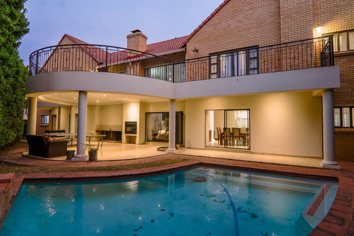 Spacious rear exterior featuring a covered patio with built-in braai, overlooking a swimming pool and a large upper-level balcony with wrought iron railings.