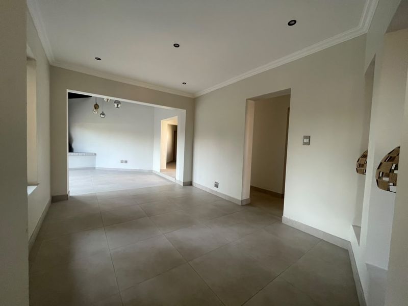 A spacious, empty living area featuring neutral tiled flooring, recessed downlighting, and coving, with an open-plan layout leading to adjacent rooms.