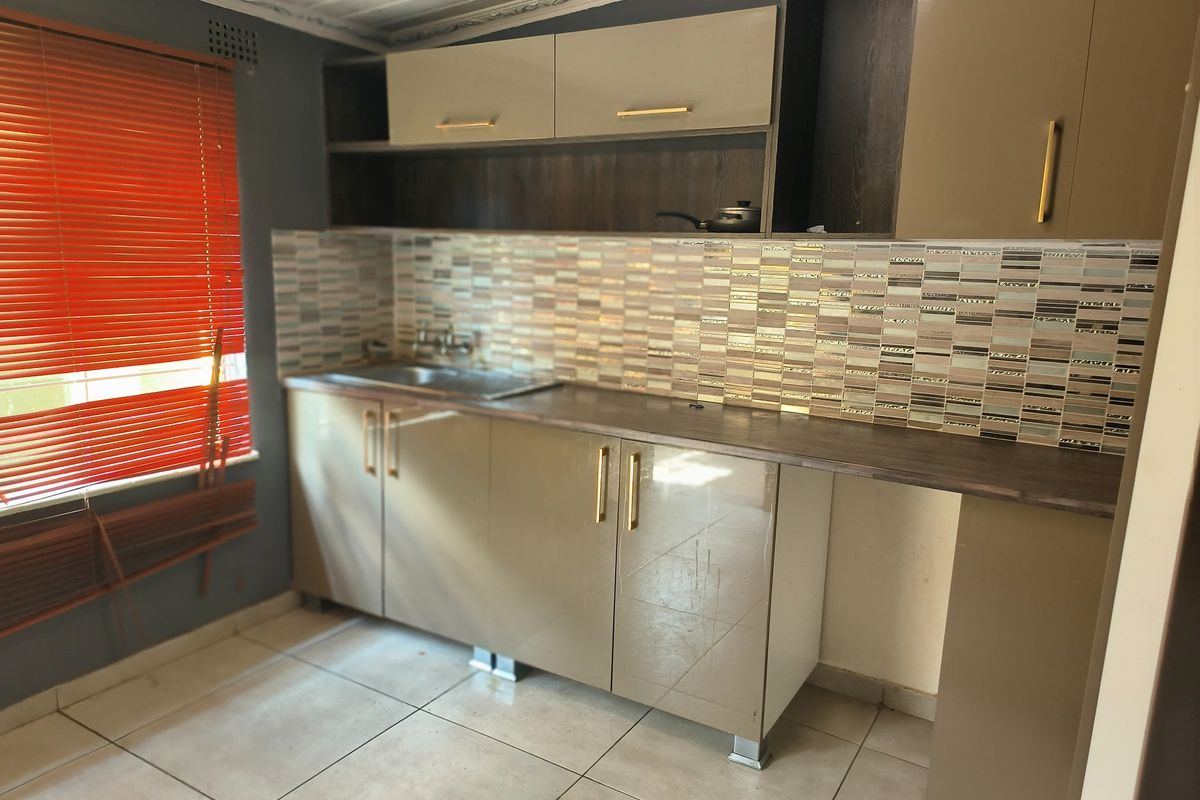 A compact kitchen featuring modern taupe cabinetry, a mosaic tiled backsplash, laminate countertops, and a stainless steel sink with tiled flooring.