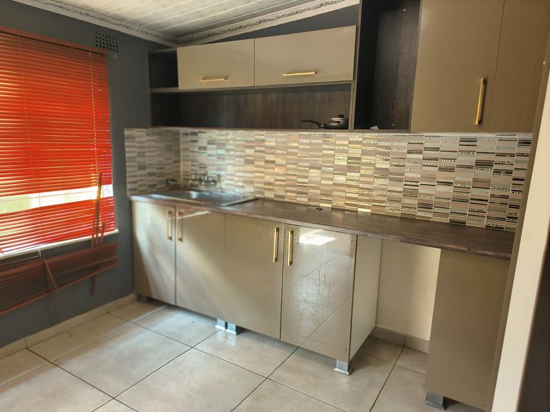 A compact kitchen featuring modern taupe cabinetry, a mosaic tiled backsplash, laminate countertops, and a stainless steel sink with tiled flooring.