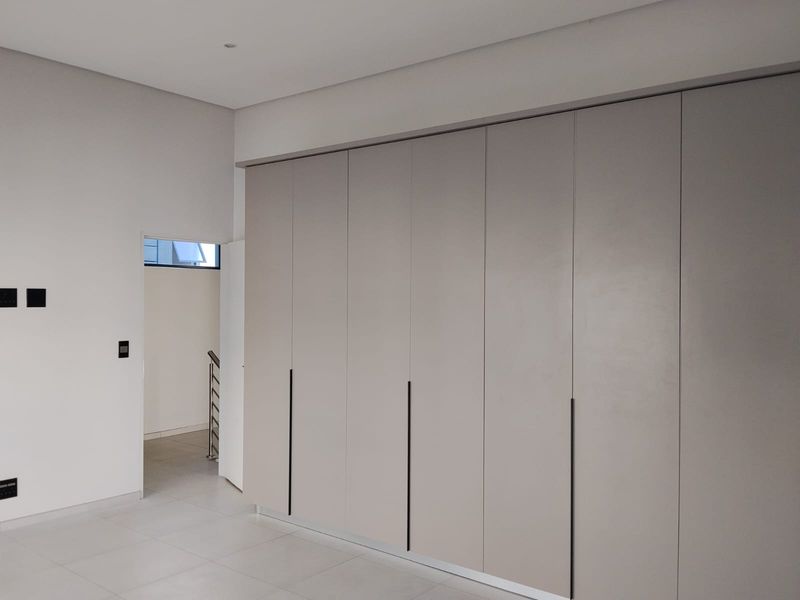 A clean, modern bedroom featuring extensive floor-to-ceiling built-in cupboards, neutral tiled flooring, and recessed ceiling lighting in a minimalist, well-maintained space.