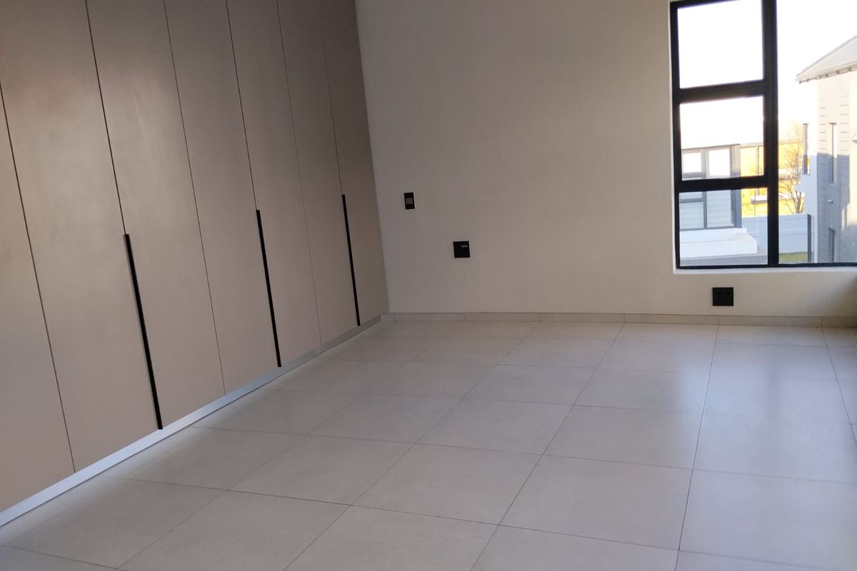 A bright, modern bedroom featuring neutral floor tiles, ample built-in cupboard storage, and large windows providing natural light.
