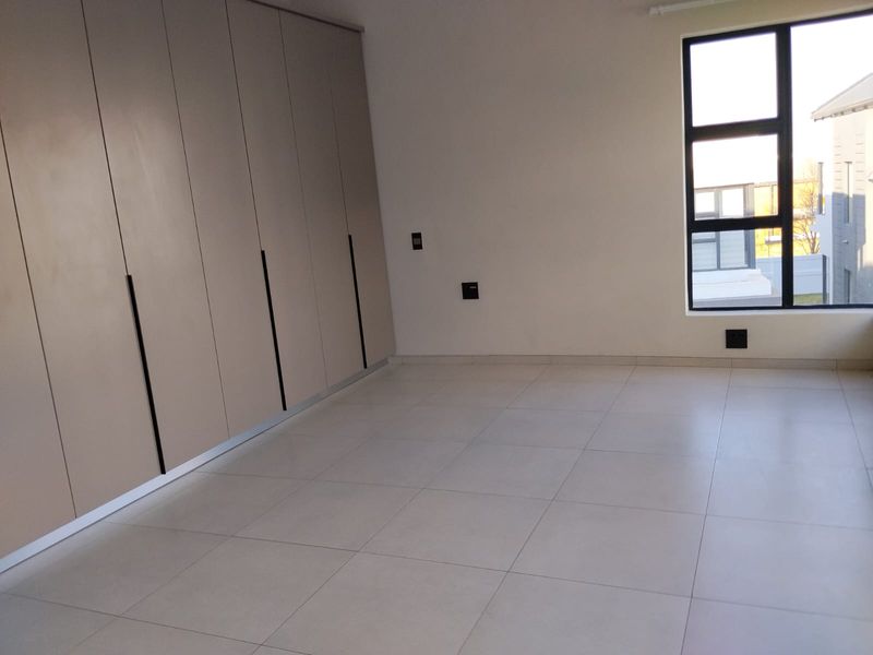 A bright, modern bedroom featuring neutral floor tiles, ample built-in cupboard storage, and large windows providing natural light.