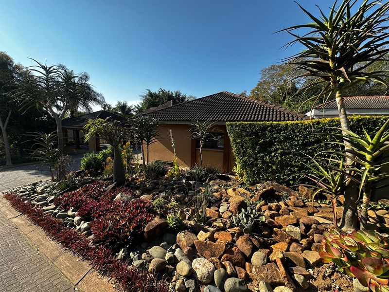 Front view of the property featuring a well-maintained rock garden, paved driveway, and established hedging under a clear blue sky.