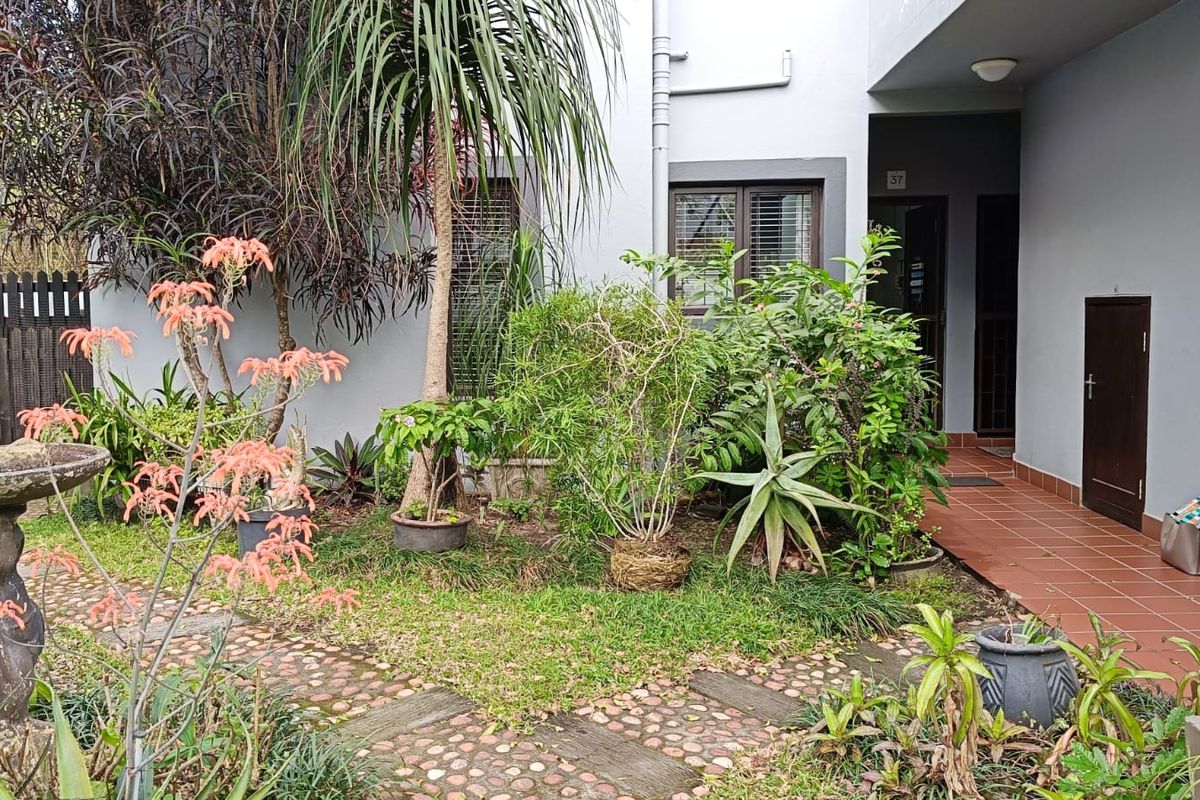 A rear garden area featuring a stone pathway, landscaped greenery, and a tiled patio leading to the apartment entrance.