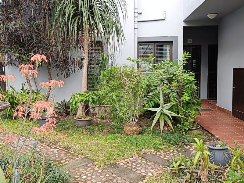 A rear garden area featuring a stone pathway, landscaped greenery, and a tiled patio leading to the apartment entrance.