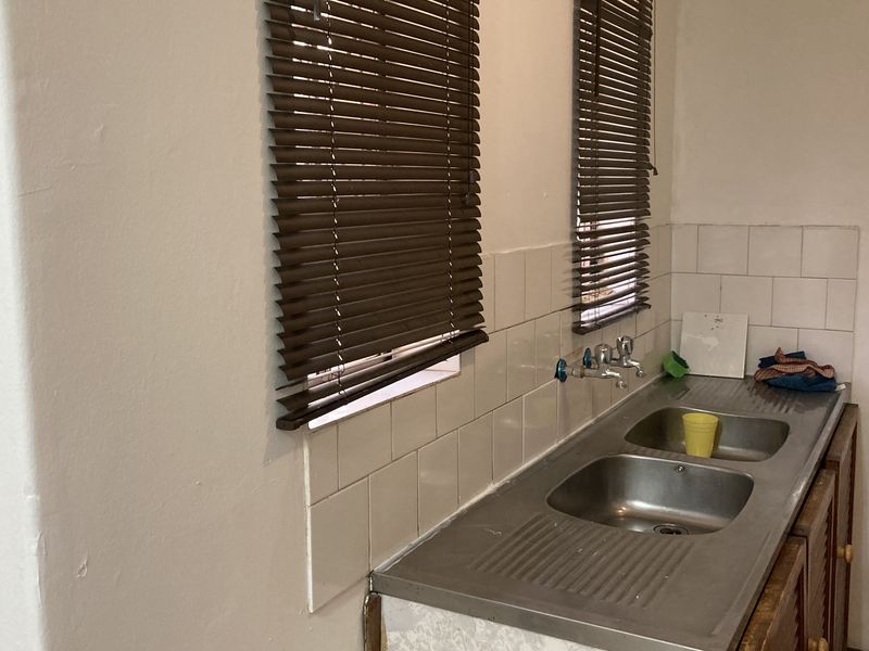 A functional kitchen area featuring a double stainless steel sink, tiled splashback, and venetian blinds, currently showing signs of age and wear.