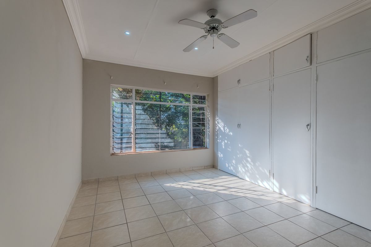 A bright, empty bedroom featuring tiled flooring, built-in cupboards, a ceiling fan, and louvered windows allowing for natural light.