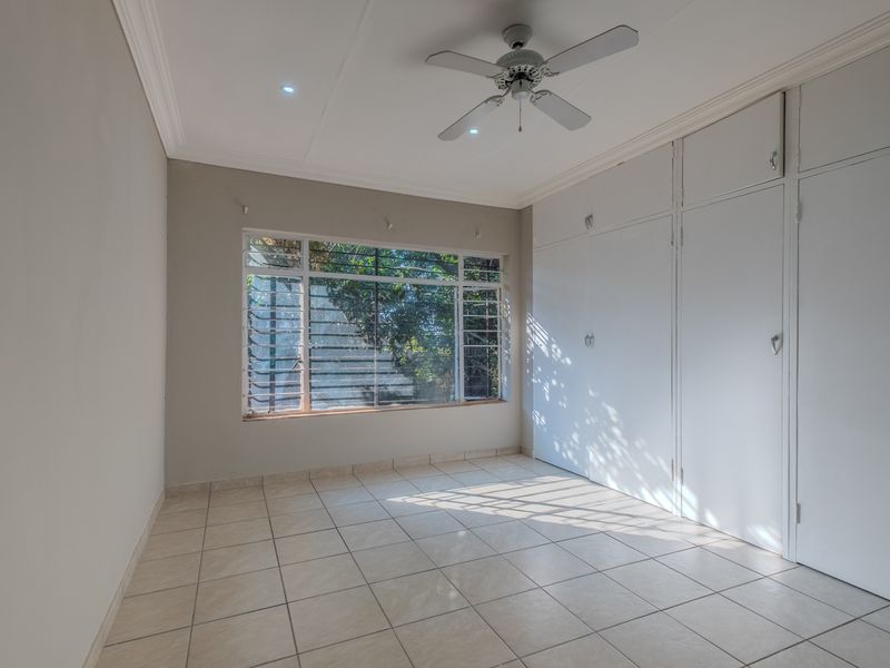 A bright, empty bedroom featuring tiled flooring, built-in cupboards, a ceiling fan, and louvered windows allowing for natural light.