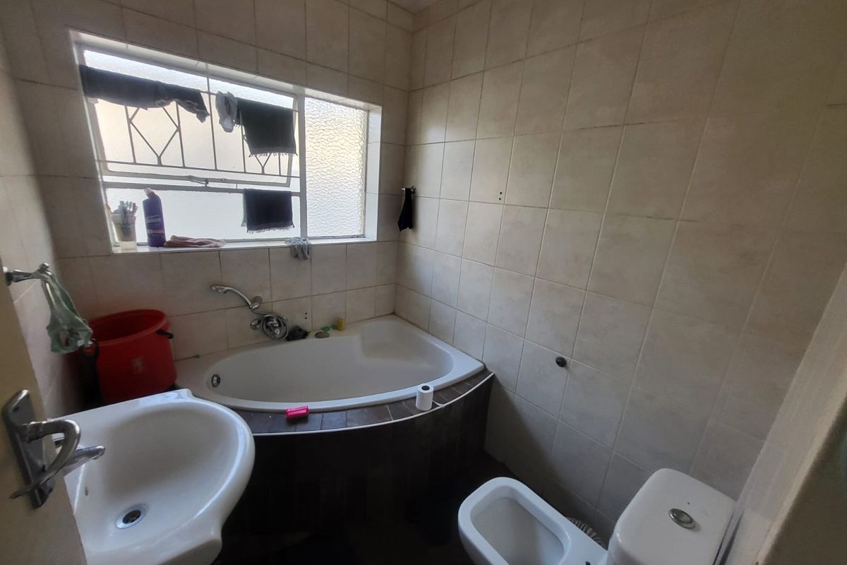 A dated bathroom featuring a built-in bathtub, pedestal basin, toilet, and bidet, finished with neutral wall tiles and security bars on the window.