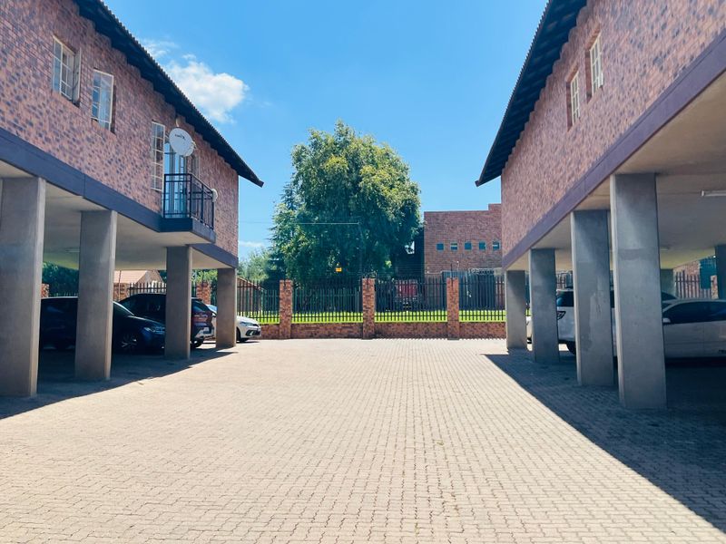 Exterior view of an apartment complex featuring a paved driveway, covered parking areas supported by concrete pillars, and brick facade construction.
