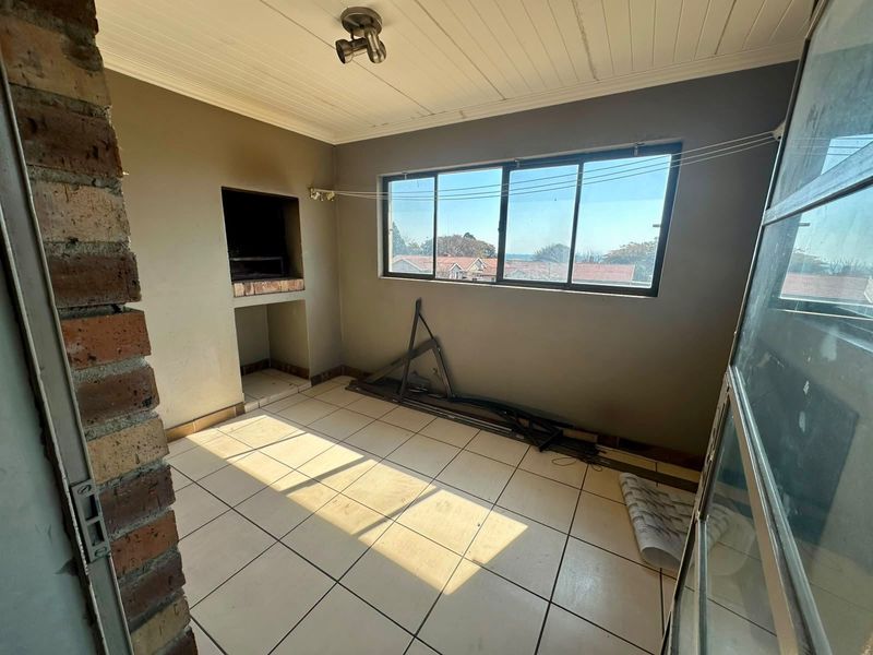 An enclosed balcony or patio area featuring a built-in braai, tiled flooring, and large sliding windows with views of the surrounding neighborhood.