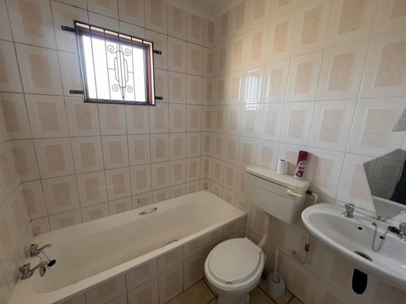 A functional bathroom featuring a bathtub, pedestal basin, and toilet, finished with dated ceramic wall tiles and fitted with security bars on the window.