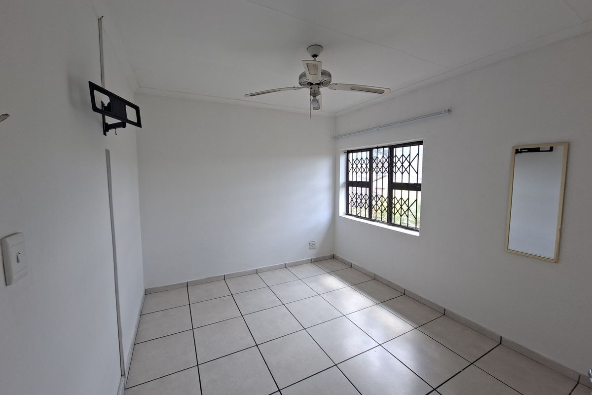 A clean, empty bedroom featuring neutral tiled flooring, a ceiling fan, and a window fitted with a security gate.