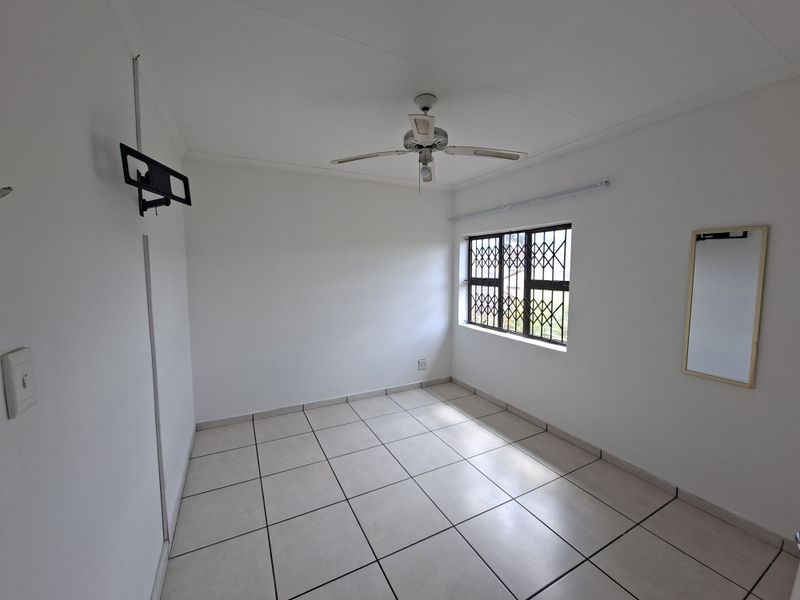 A clean, empty bedroom featuring neutral tiled flooring, a ceiling fan, and a window fitted with a security gate.