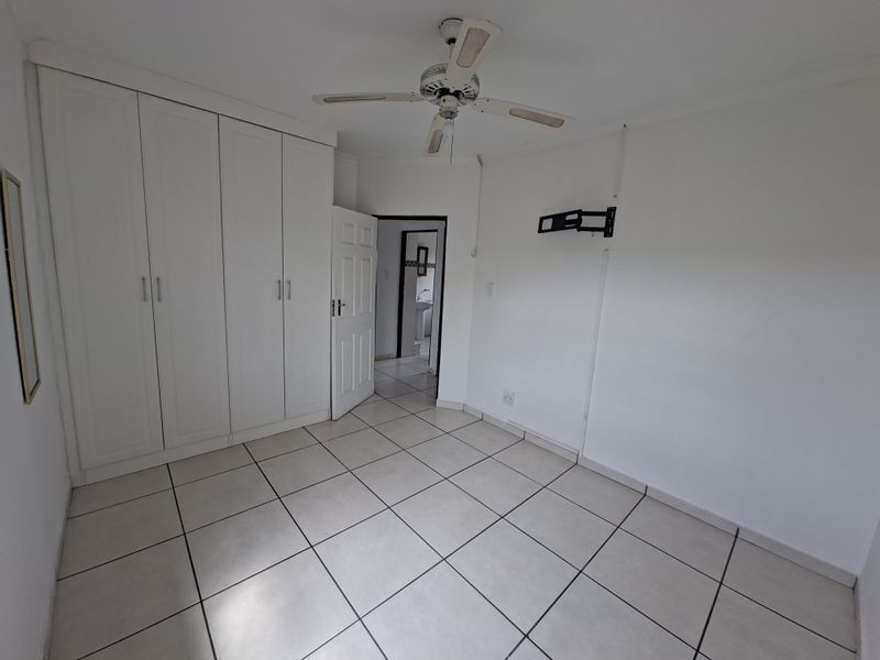 A clean, empty bedroom featuring neutral tiled flooring, built-in cupboards, a ceiling fan, and a wall-mounted TV bracket, offering a versatile space.