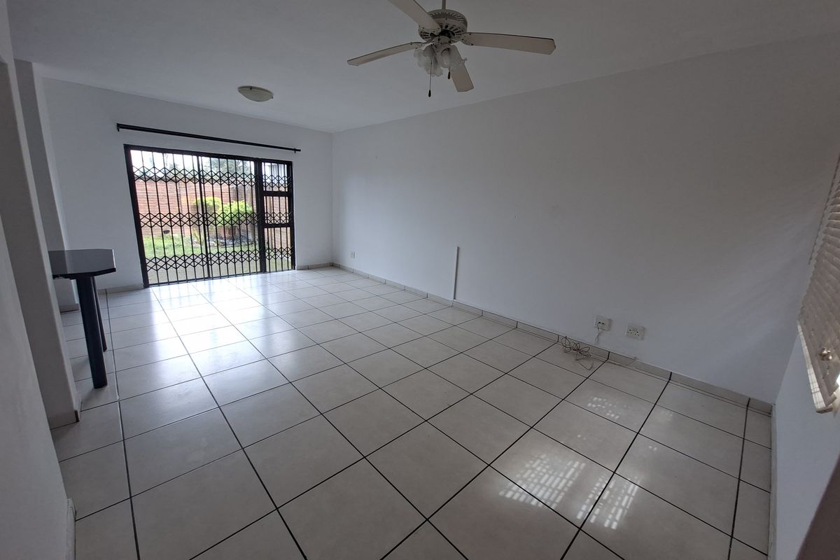 An empty, open-plan living area featuring neutral tiled flooring, a ceiling fan, and a sliding door with a security gate leading to the exterior.