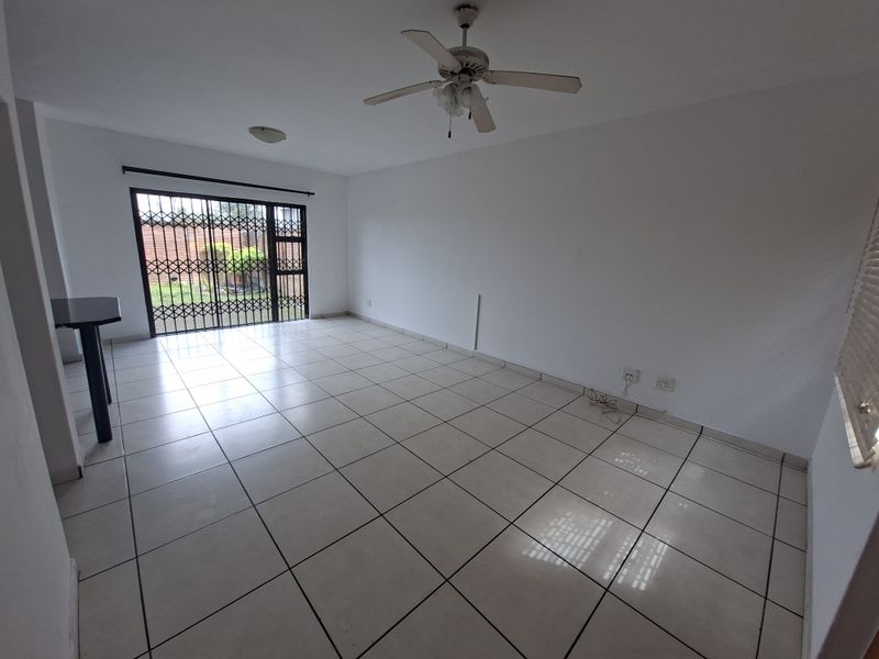An empty, open-plan living area featuring neutral tiled flooring, a ceiling fan, and a sliding door with a security gate leading to the exterior.