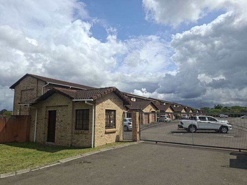 Front view of a townhouse complex featuring a brick facade, tiled roofing, and a secure gated entrance with paved driveway access.