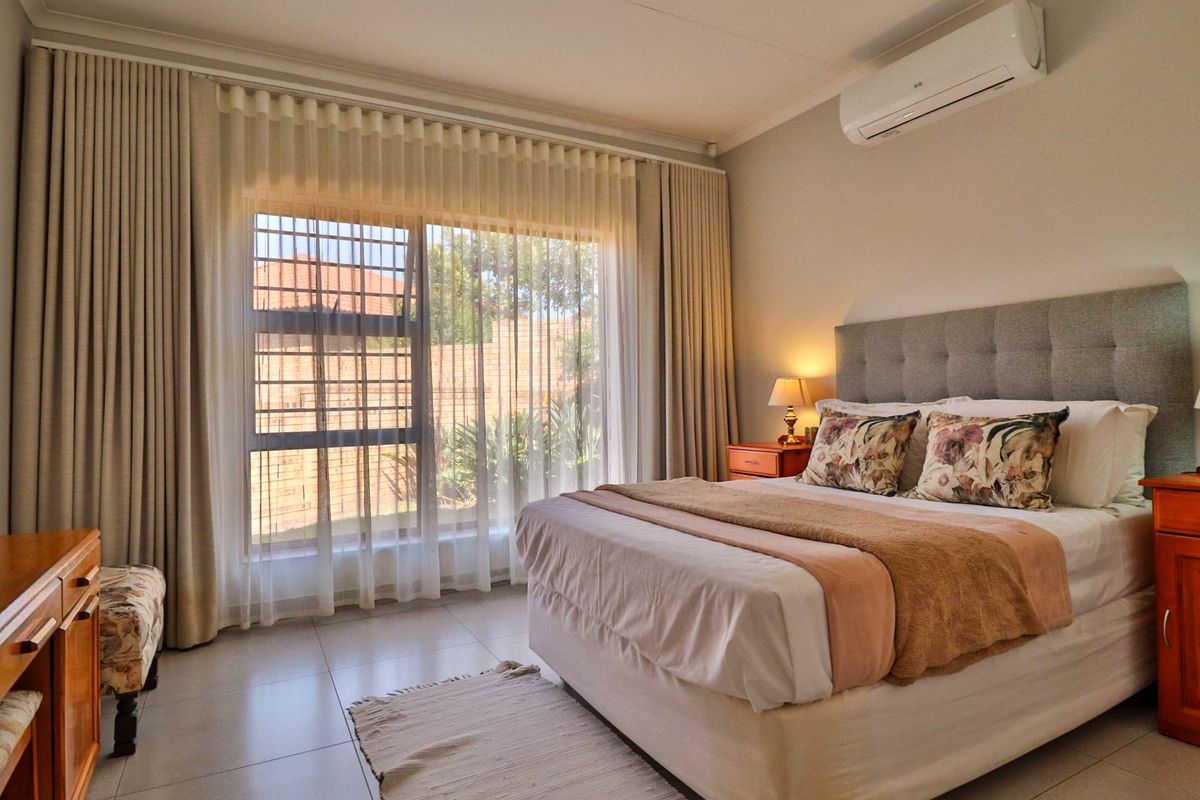 A well-maintained bedroom featuring tiled flooring, a wall-mounted split-system air conditioner, and large windows with security bars and floor-to-ceiling curtains.