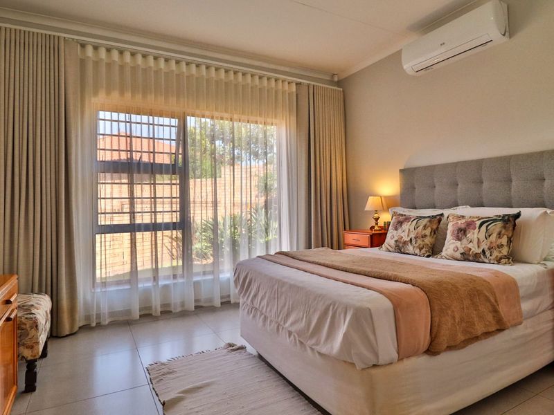 A well-maintained bedroom featuring tiled flooring, a wall-mounted split-system air conditioner, and large windows with security bars and floor-to-ceiling curtains.