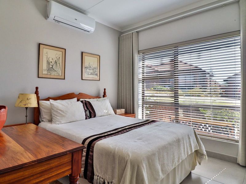 A well-maintained bedroom featuring tiled flooring, a wall-mounted split-system air conditioner, and large windows fitted with venetian blinds and curtains.