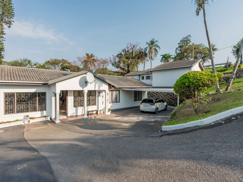 Front exterior view of a townhouse featuring a tiled roof, covered carport, and paved driveway with established garden surroundings.