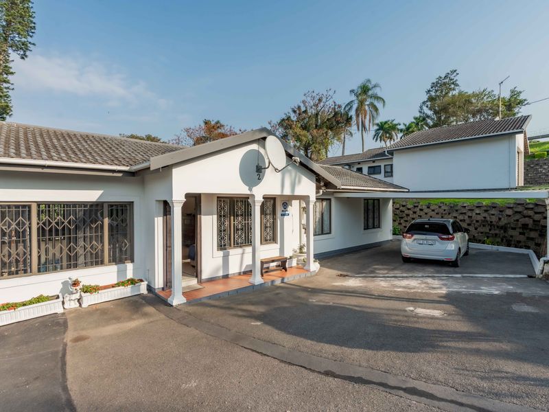 Front elevation of a townhouse featuring a covered entrance porch, decorative columns, security bars on windows, and an asphalt driveway with a retaining wall.