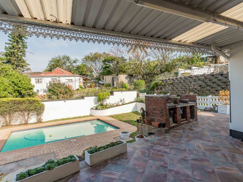 Covered outdoor patio featuring a built-in brick braai, overlooking a private swimming pool and landscaped garden area.