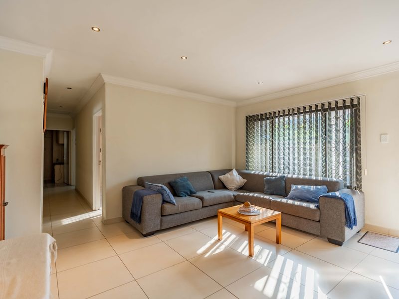 A bright and spacious living room featuring neutral tiled flooring, recessed downlighting, and large windows with vertical blinds, providing a comfortable and well-lit living space.