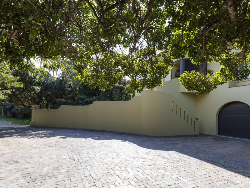 A well-maintained front exterior featuring a paved driveway, a curved retaining wall, and a secure garage entrance shaded by mature trees.