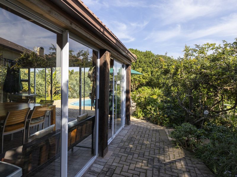 A covered patio area featuring sliding glass doors, timber support posts, and paved flooring, overlooking a private garden and swimming pool.