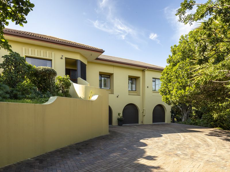 Front elevation of a yellow house featuring a paved driveway, retaining wall, and double garage with arched entrances.