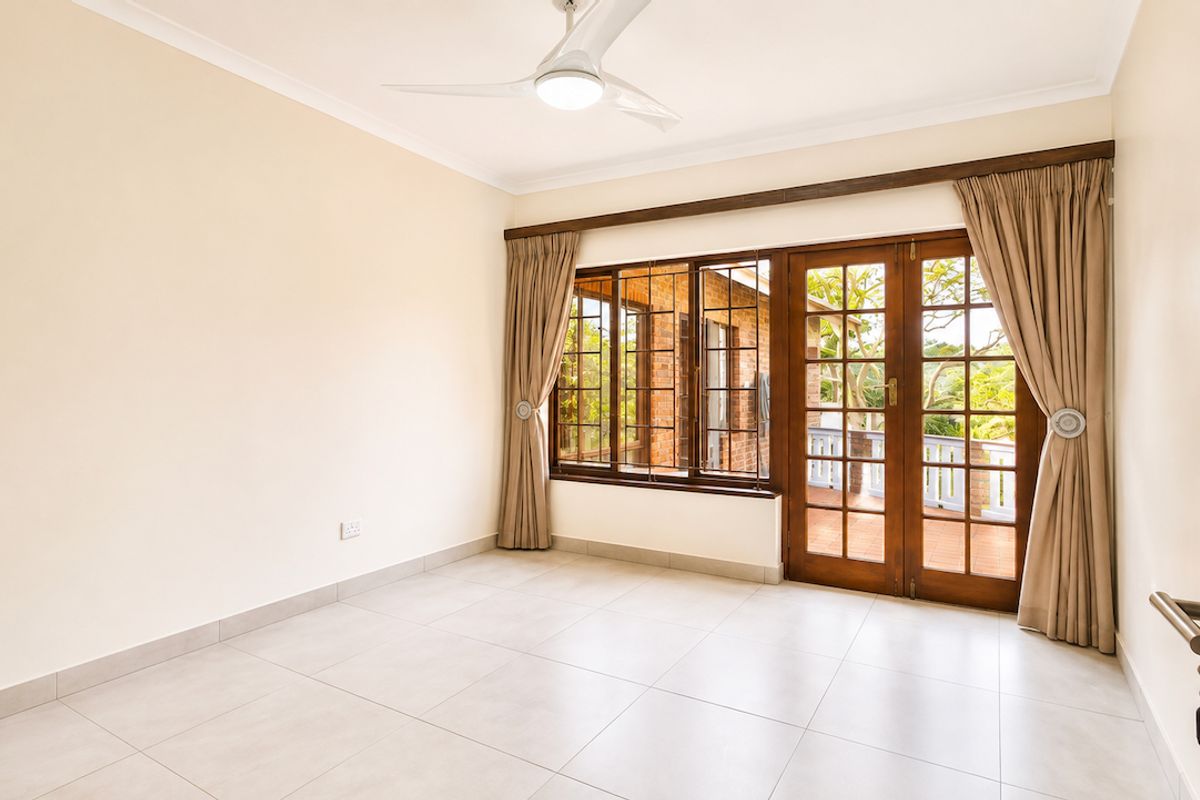 A bright, empty bedroom featuring neutral tiled flooring, a ceiling fan, and wooden french doors providing access to an outdoor patio area.