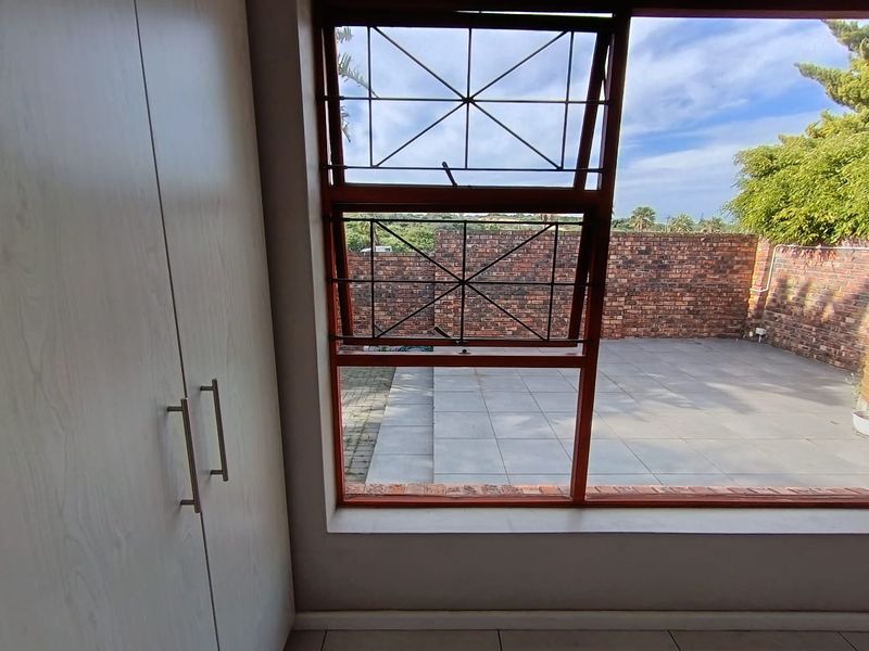 A bright bedroom featuring built-in cupboards and a large window with security bars, offering a view of the paved rear patio area.