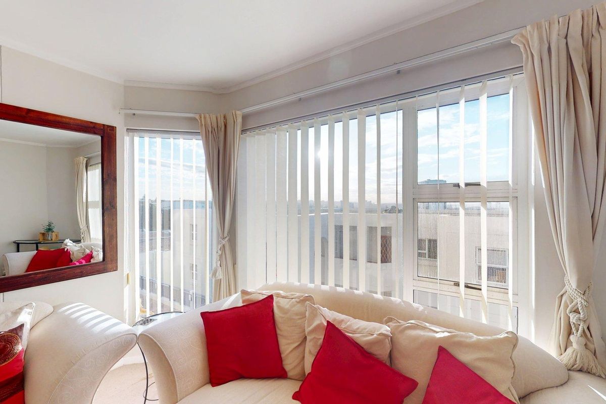 A bright living room area featuring a large bay window with vertical blinds and curtain tracks, offering natural light and an urban view.